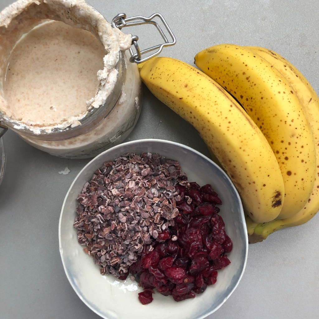 A pot of sourdough starter with some ripe bananas on a kitchen top next to cacao nibs and dried cranberries in a handmade bowl