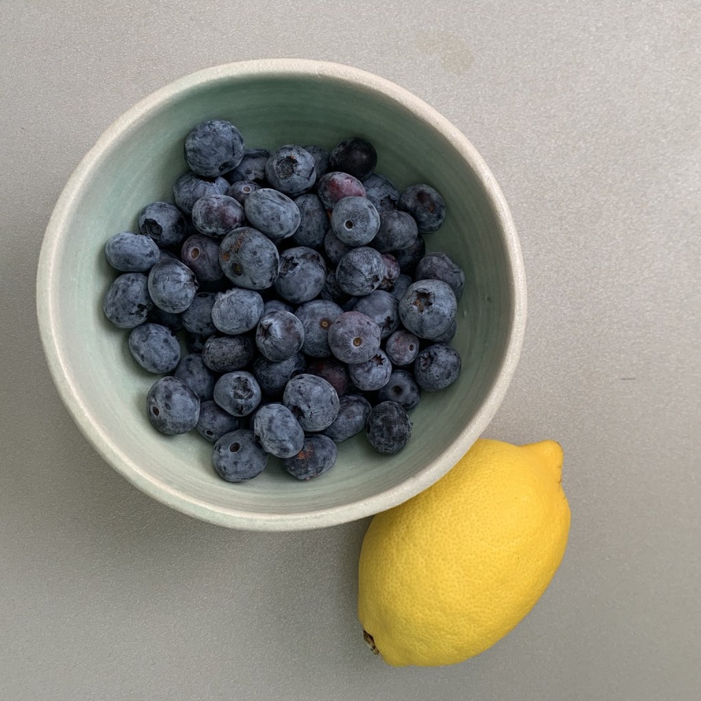 A handmade ceramic bowl filled with blueberries next to a wole fresh lemon